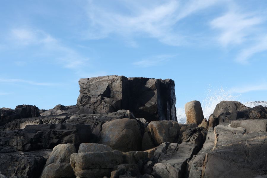 A monolithic block of stone stands against the sky as spray bursts behind it